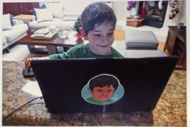 Elliot as a child, around the age of 7, sitting in front of his first laptop computer, smiling.